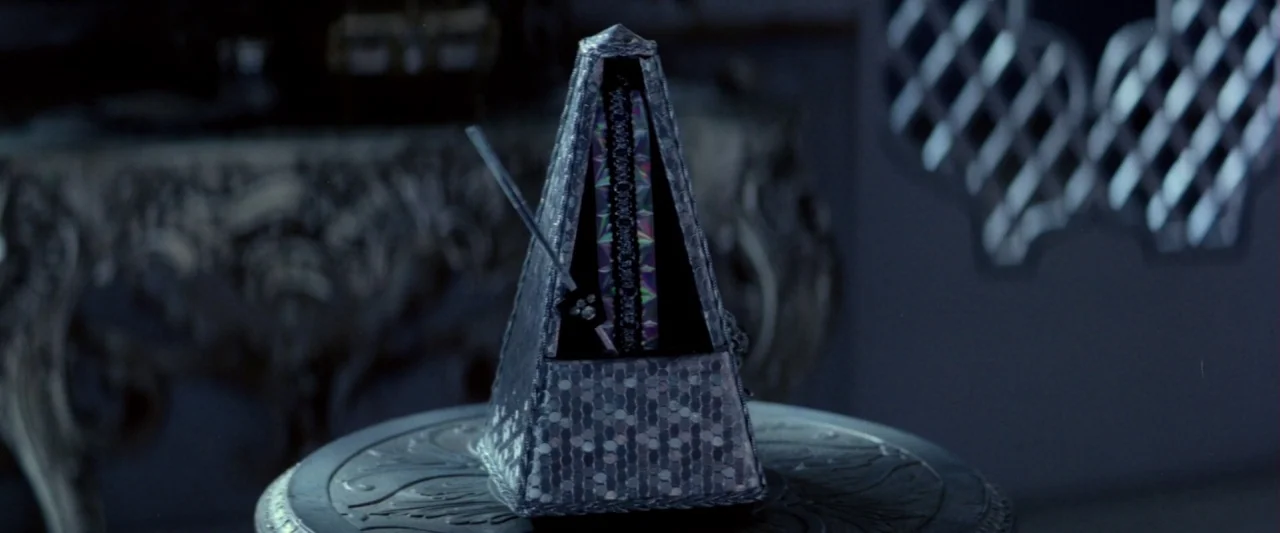 Lisztomania (1975) movie still: closeup — A decorative pyramidal metronome sits centered on a small ornate table in a dark room. The object features…