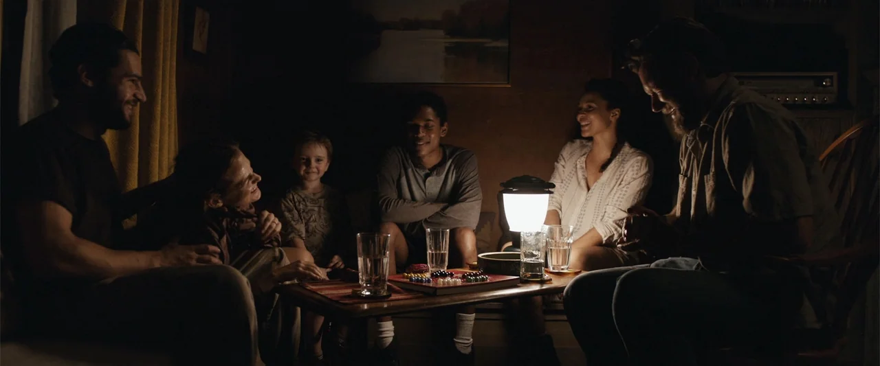 It Comes at Night (2017) movie still: medium — A dimly lit room at night features six people gathered around a small table. The group plays a board…