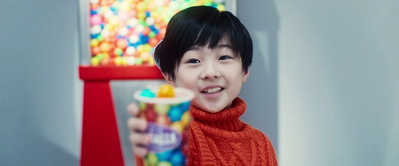 Prisoners of the Ghostland (2021) movie still: closeup — An interior space features a red gumball dispenser in the blurred background. A young boy…