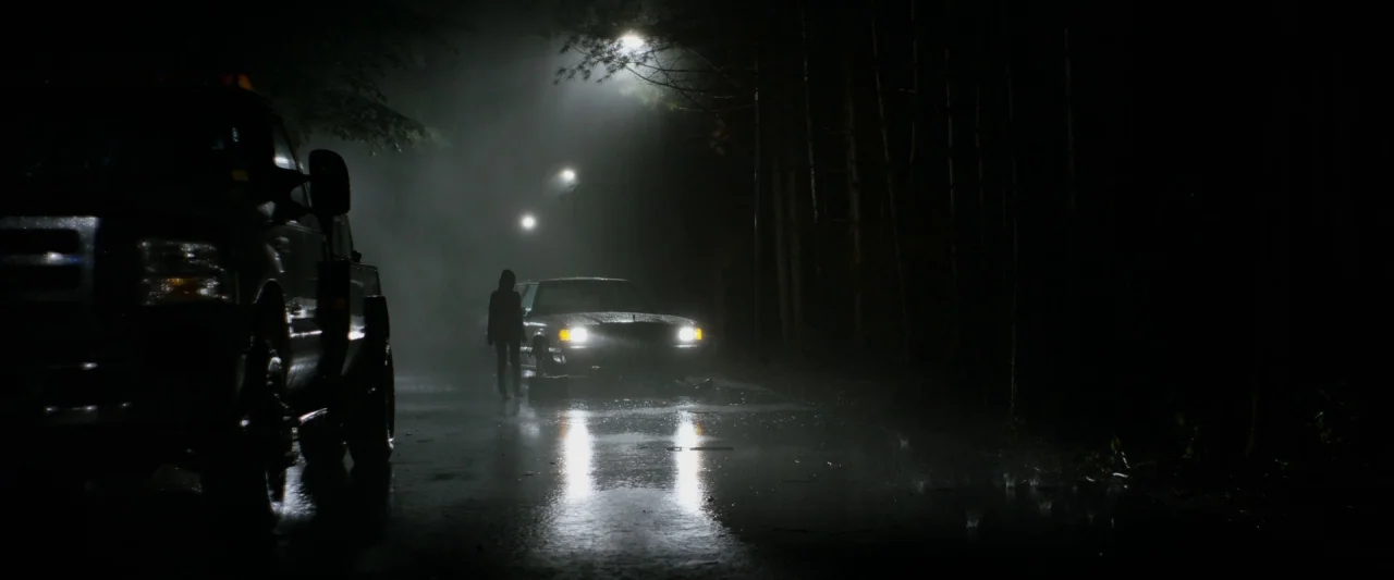 The Monster (2016) movie still: wide — A dark, rain-slicked asphalt road runs through a forested area under night sky. A person stands facing a…