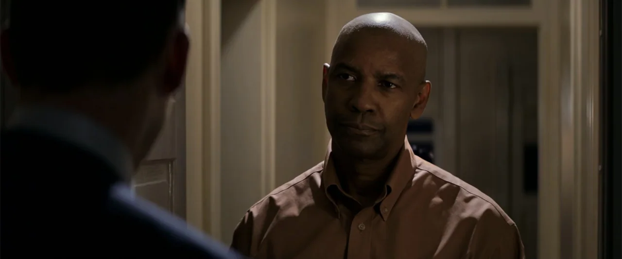 The Equalizer (2014) movie still: medium — A dimly lit indoor hallway with light-colored walls and doors. A man stands in the foreground looking at…