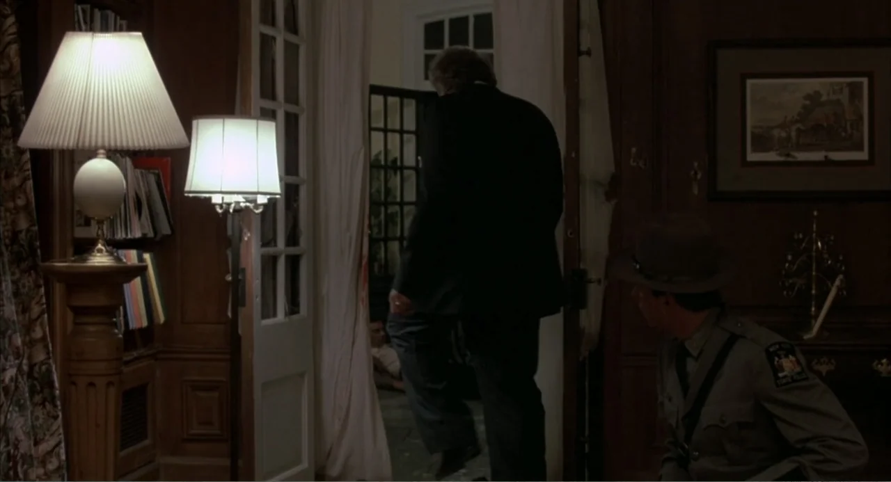 F/X (1986) movie still: medium — A dimly lit paneled room with bookshelves and a framed picture on the wall. A man in a suit steps through a doorway…