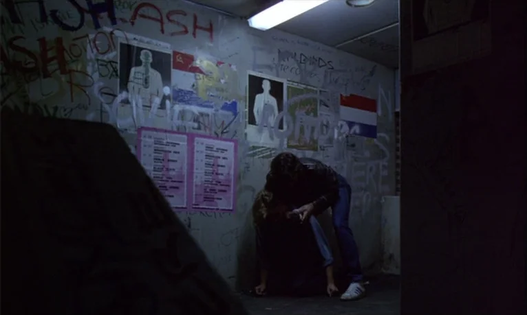 Spetters (1980) movie still: medium — A dimly lit room features walls covered in graffiti and posters depicting human figures. One individual stands…