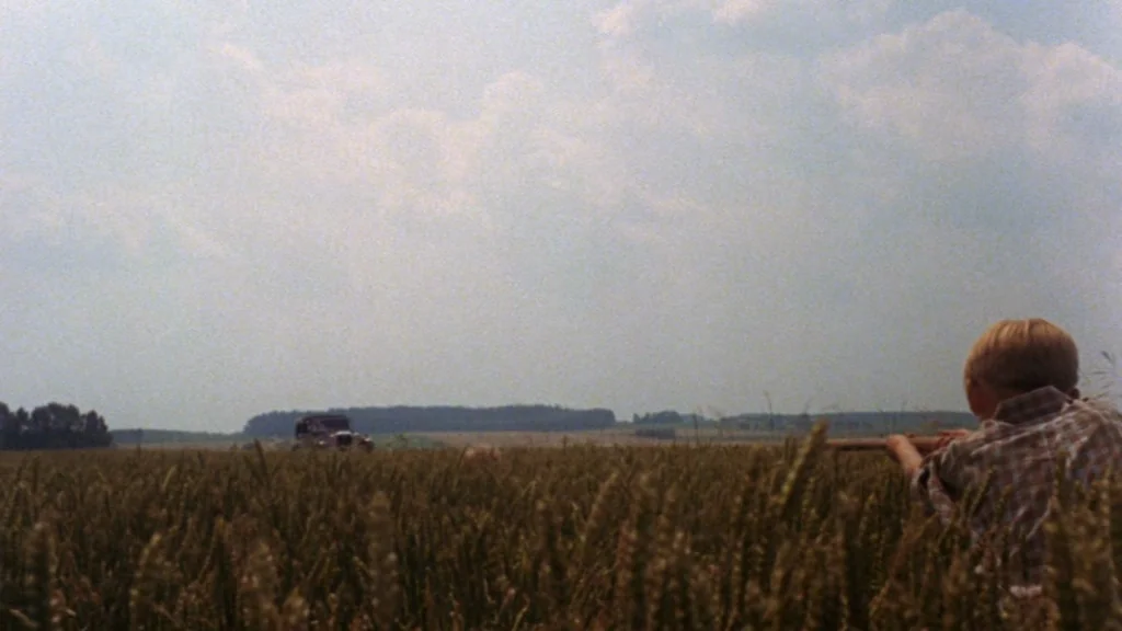 Cabaret (1972) movie still: wide — A vast, open wheat field stretches toward a distant line of trees under sky. A young boy holding a rifle aims…