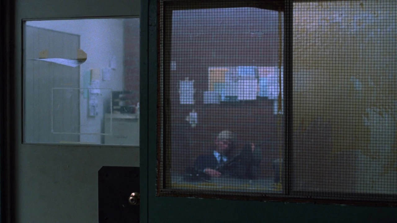 Repo Man (1984) movie still: medium — A dark security booth features a metal-framed glass partition with wire-mesh texture. A man sits at a desk…