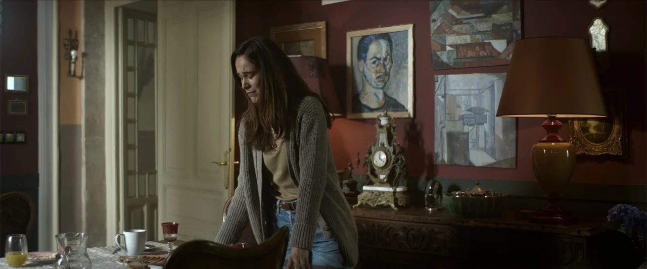 Tell Me Softly (2025) movie still: medium — A domestic room features dark burgundy walls decorated with several framed paintings and a clock. A…