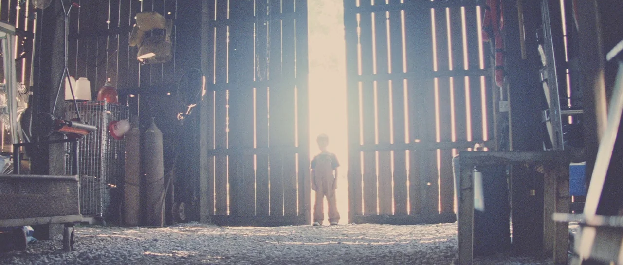 Looper (2012) movie still: wide — A dimly lit barn interior contains various tools and metal containers on the sides. A young boy stands centrally…