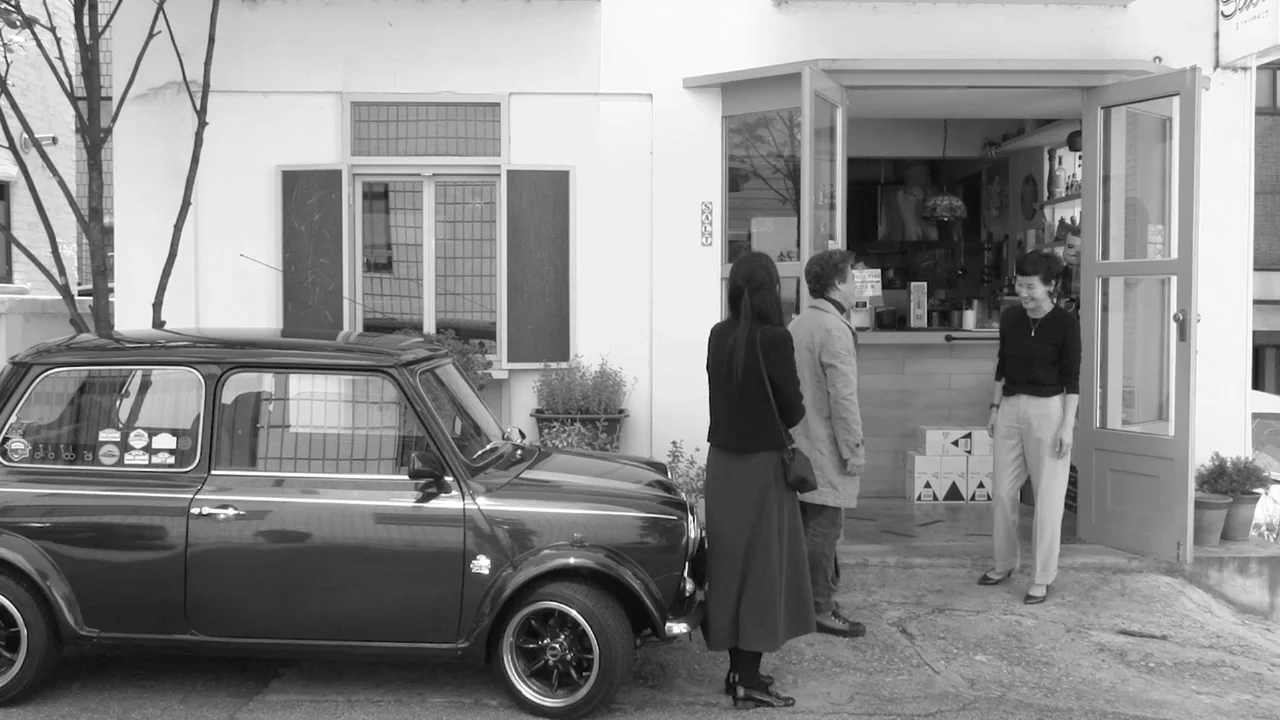 Walk Up (2022) movie still: fullbody — A white building with an open door stands behind a parked black mini car. Three people stand outside the…