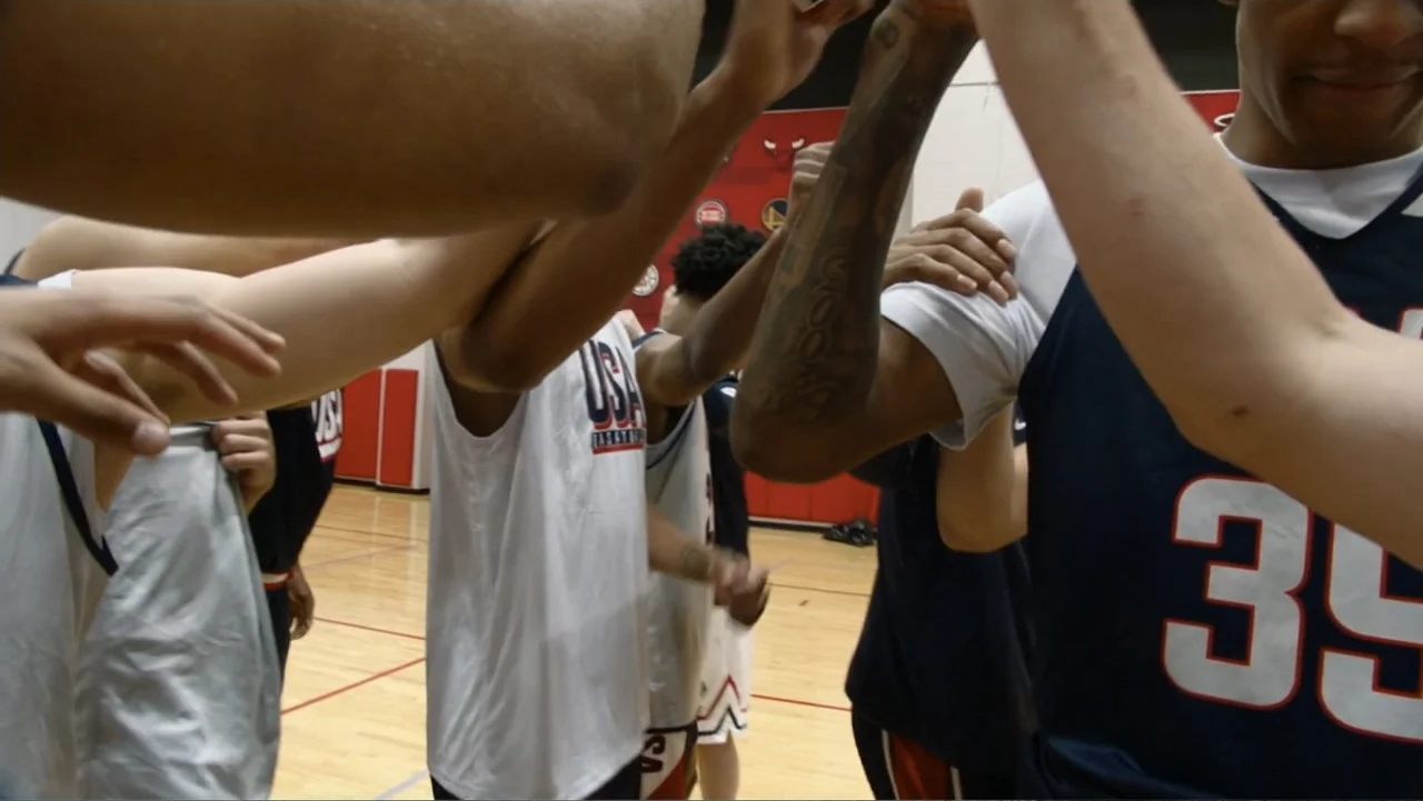 We Beat the Dream Team (2025) movie still: medium — A group of basketball players huddle together on a hardwood court in front of a red wall. Their…