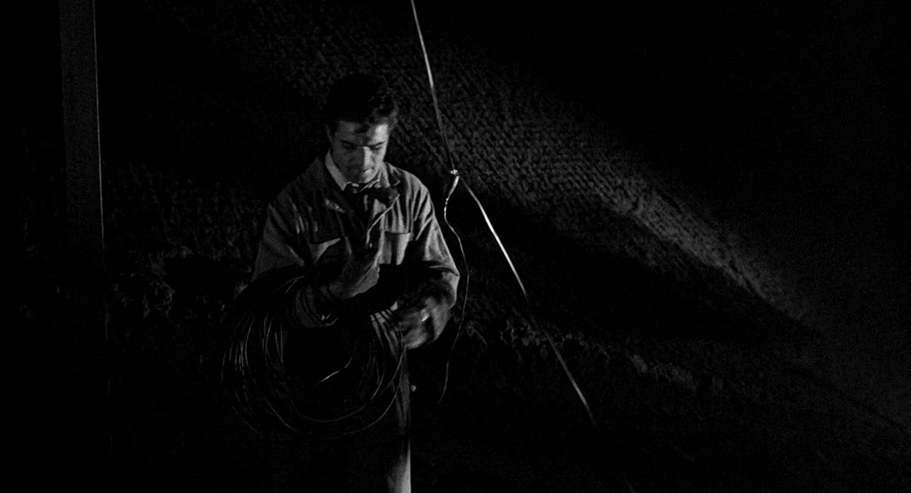 Murder By Contract (1958) movie still: medium — A man stands in an unidentifiable, dark outdoor space at night. He is holding a large coil of thick…