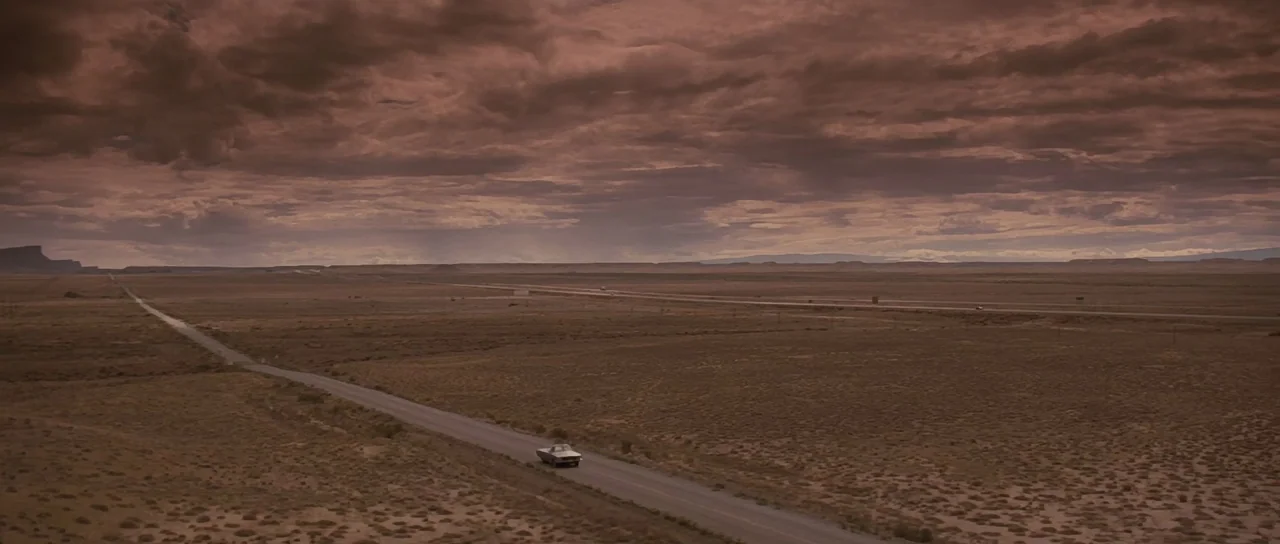 Thelma & Louise (1991) movie still: establishing — A vast, flat desert landscape extends to the horizon under a dark, overcast sky. A lone car…