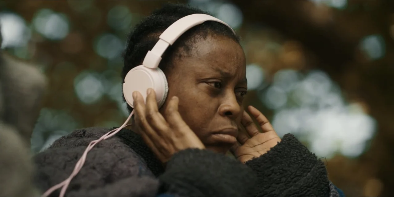 Autumn Beat (2022) movie still: closeup — A woman sits outdoors with blurred trees in the background. She wears light-colored headphones while…