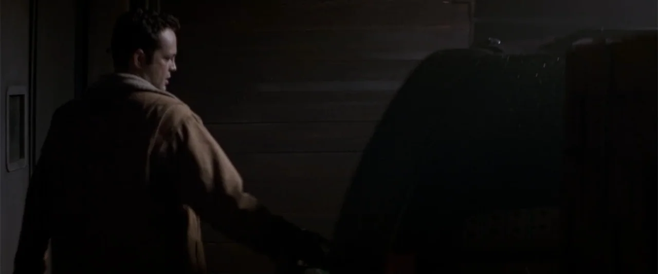 Angst (2017) movie still: medium — A dark industrial workspace with wooden wall paneling and dim ambient light. A man in a brown jacket stands near…