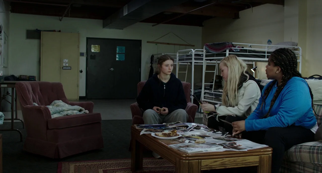 Leave No Trace (2018) movie still: wide — A shared living area features bunk beds, a coffee table, and upholstered furniture. Three young women sit…
