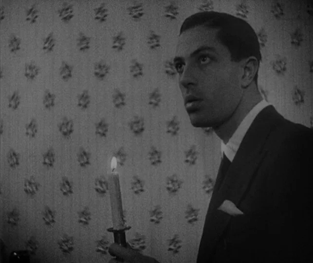 Vampyr (1932) movie still: medium — An interior space features floral patterned wallpaper covering the walls. A man in a suit stands holding a lit…