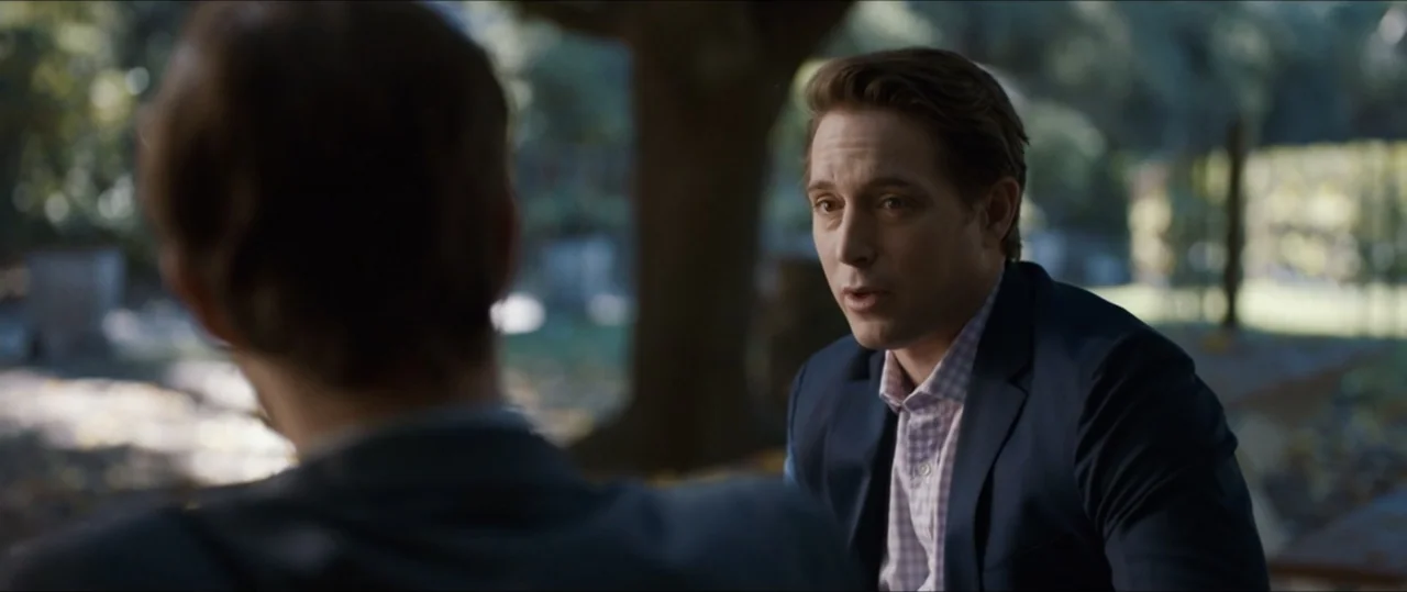 Plus One (2019) movie still: medium — An outdoor park setting features mature trees and dappled sunlight in the background. A man in a blue blazer…