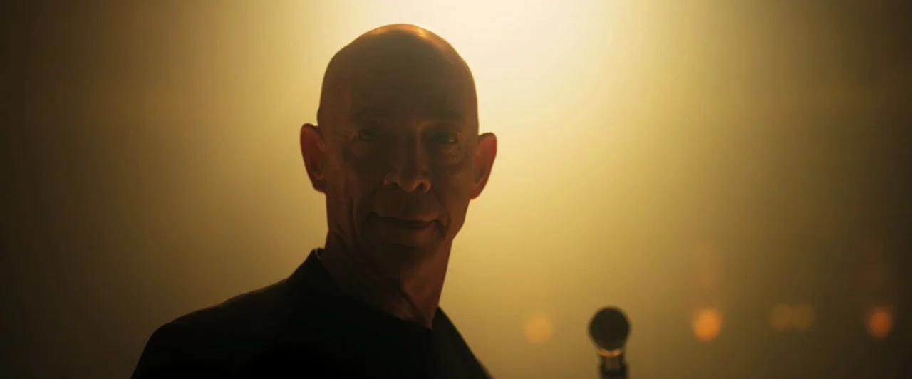 Whiplash (2014) movie still: closeup — A dimly lit room with a bright warm light source behind a man. A bald man faces the camera with a microphone…