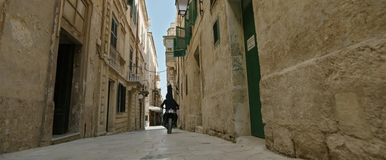 Hounds of War (2024) movie still: wide — A narrow stone street features aged limestone buildings with green shutters and brown doors. A person…