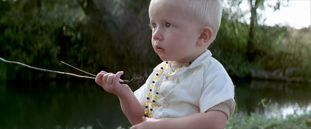 Silent Light (2007) movie still: medium — A toddler stands near a dark, murky river lined with dense green foliage. The child holds a thin twig…