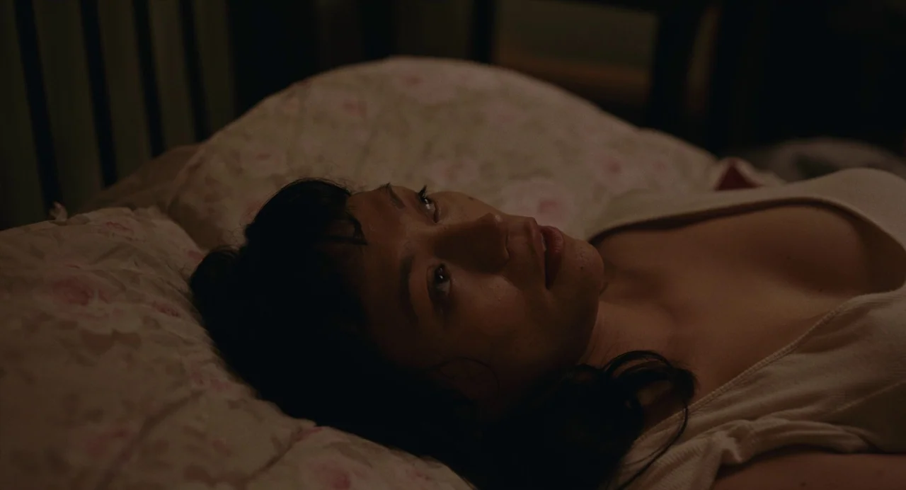 Lingua Franca (2019) movie still: closeup — A dark bedroom with floral patterned bedding illuminated by minimal light. A young woman lies…