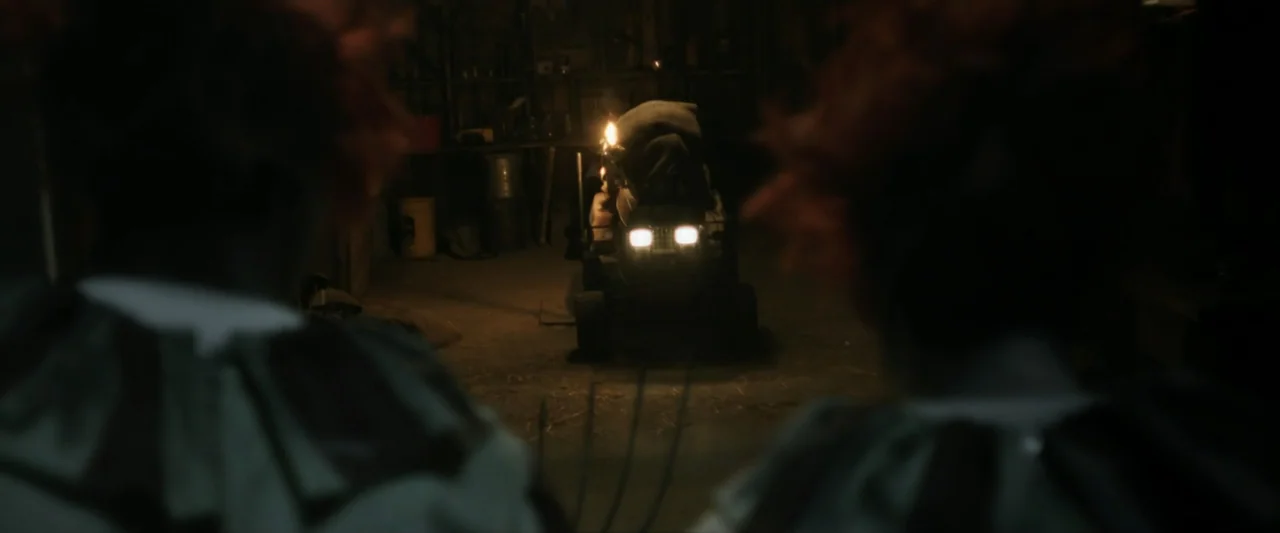 Clown in a Cornfield (2025) movie still: wide — A dark, cluttered garage interior is illuminated only by the headlights of a machine. Two blurry…