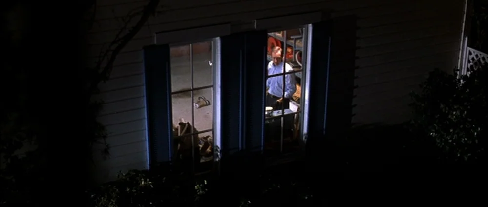 American Beauty (1999) movie still: wide — The exterior of a two-story home at night featuring dual white-framed windows. A man stands behind the…