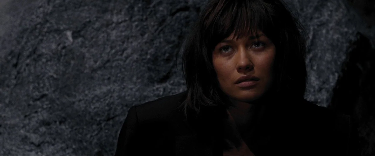 Quantum of Solace (2008) movie still: closeup — A dark cave interior with rough, gray rock textures behind the subject. A woman with dark hair…