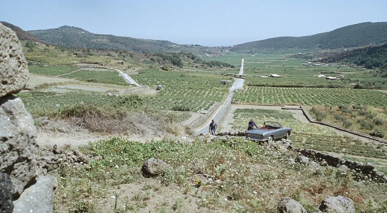 A Bigger Splash (2015) movie still: wide — A rural landscape features sprawling terraced vineyards on rolling hills under a clear sky. A grey…