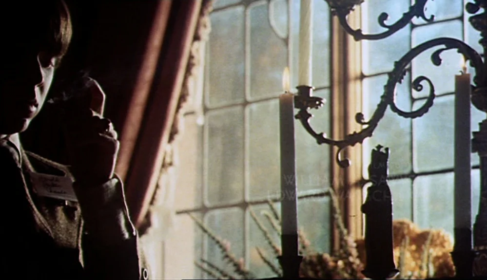 Harold and Maude (1971) movie still: medium — A dimly lit room features a leaded glass window and a ornate metal candelabra. A young male in a…