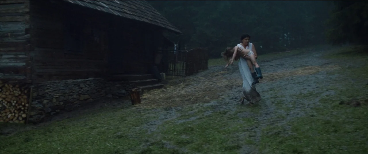 Nightsiren (2022) movie still: wide — A rural setting features a dark wooden cabin beside a damp, muddy field. A woman in a white dress walks across…