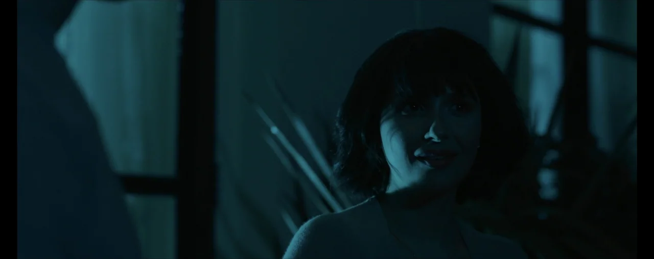 The Wrong Tenant (2022) movie still: closeup — A dimly lit room features a woman partially illuminated by blue light near foliage. The woman looks…