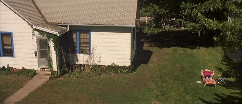 The Straight Story (1999) movie still: wide — A white house with blue trim sits adjacent to a grassy suburban backyard. An elderly woman lies on a…