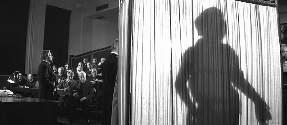 The Elephant Man (1980) movie still: medium — A dark lecture hall with tiered wooden seating containing a seated audience. A man stands near a table…
