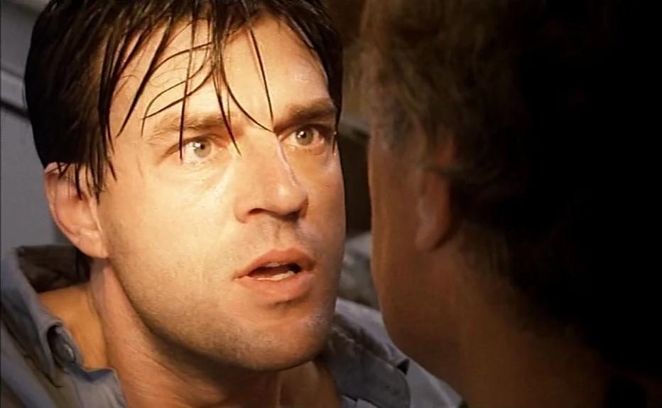 The Fourth Man (1983) movie still: closeup — Two individuals engage in a conversation within a dimly lit interior space. One man stares ahead with a…