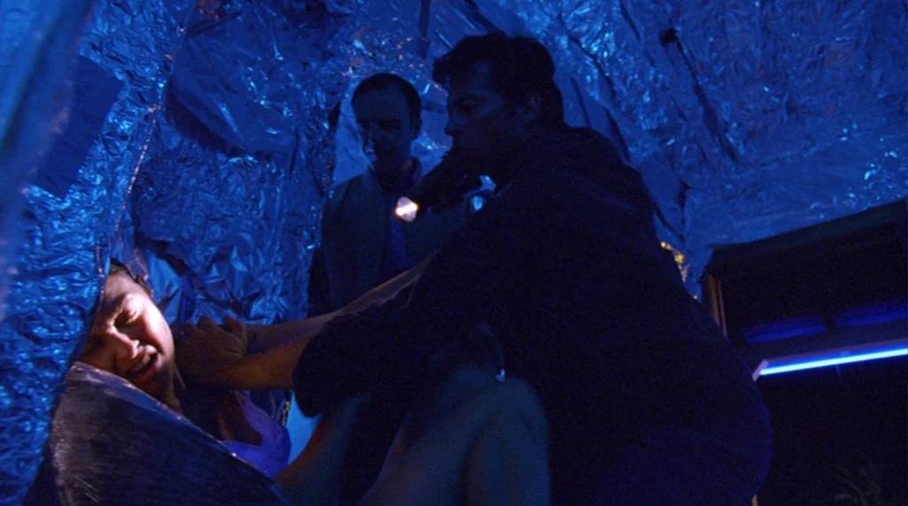 Bug movie still: medium — A confined space lined with crinkled aluminum foil reflecting intense blue light. A man aims a flashlight at a woman lying…
