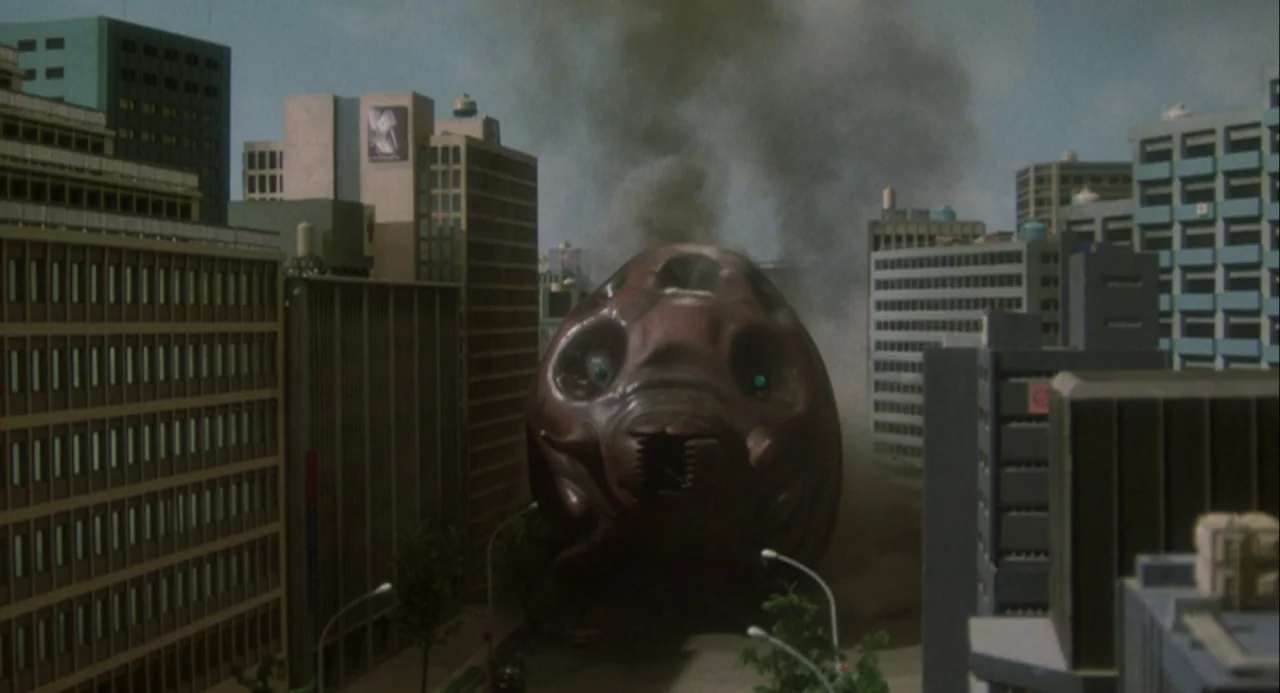 Godzilla and Mothra: The Battle for Earth (1992) movie still: wide — A dark, bulbous monster stands between several tall, model-like office…