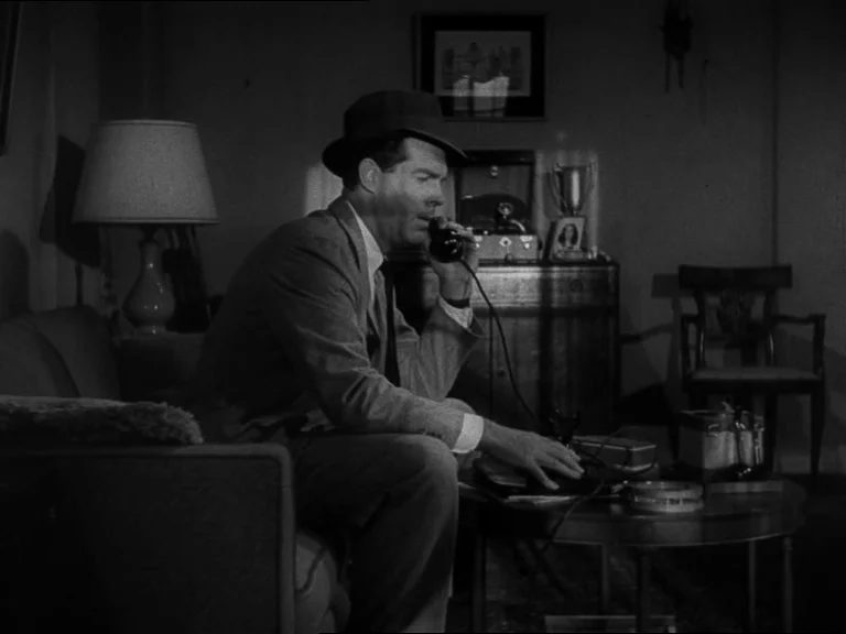 Double Indemnity (1944) movie still: medium — A dimly lit room features a sofa and a coffee table with various items. A man sits on the couch while…