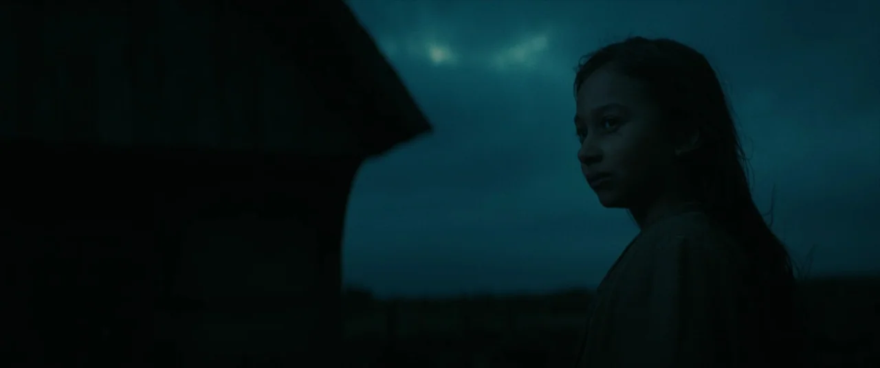 The Promised Land (2023) movie still: medium — A dark, open field under a dim blue night sky features a wooden structure. A young girl stands in the…
