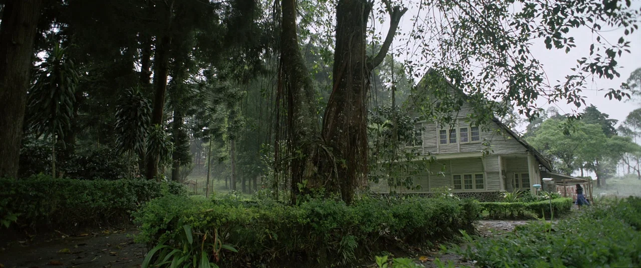 Satan’s Slaves (2017) movie still: wide — A wooden structure with a gabled roof stands nestled within a dense forest clearing. Tall trees and…