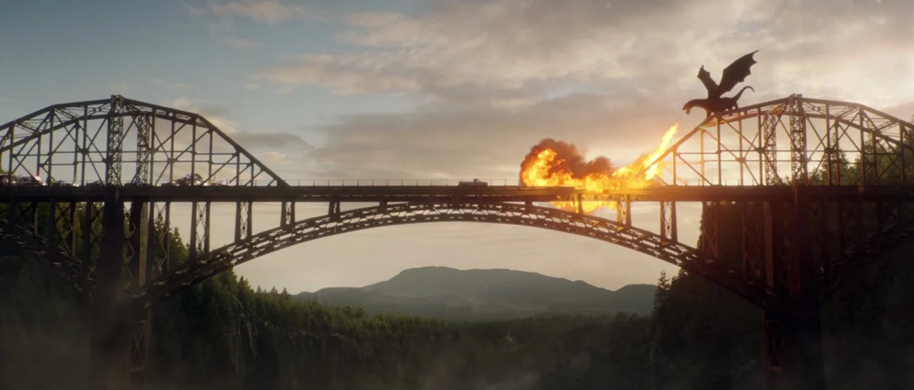 Pete’s Dragon (2016) movie still: wide — A large steel truss bridge spans a deep, forested valley under a cloudy sky. A winged dragon perches on the…