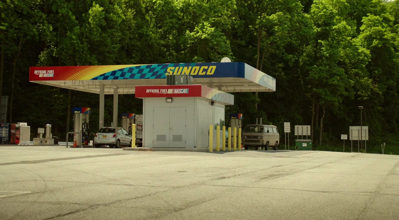 Wiener-Dog (2016) movie still: establishing — A Sunoco gas station sits at the base of a dense, green deciduous forest. Two vehicles are parked near…