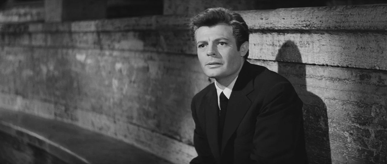 La Dolce Vita (1960) movie still: medium — A man sits against a textured stone wall during a night scene. The man looks toward the left of the frame…