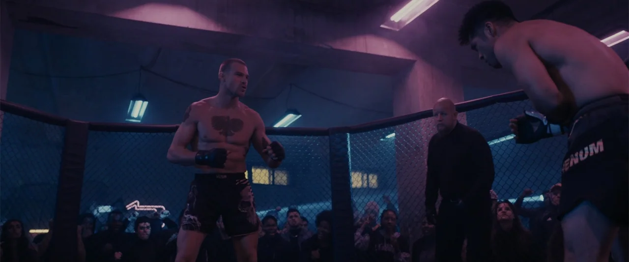Perfect Addiction (2023) movie still: medium — A dimly lit industrial fighting ring is surrounded by a wire mesh fence. Two shirtless muscular…