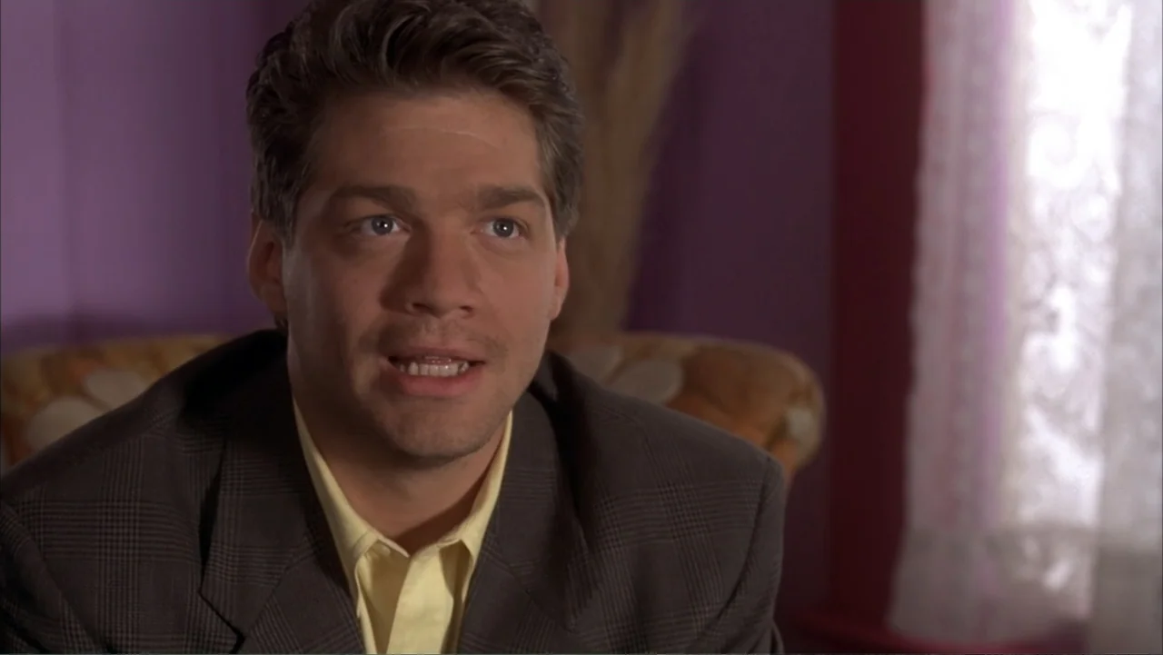 The Wedding Singer (1998) movie still: medium — A man sits in a room with purple walls and a patterned chair. The man talks toward another person…