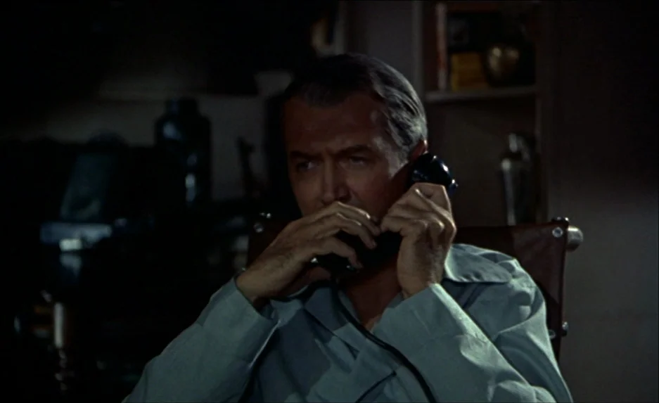 Rear Window (1954) movie still: medium — A dark apartment interior featuring a man seated in shadow. He is holding a telephone receiver to his ear…