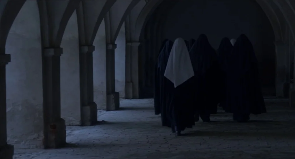 The Innocents (2016) movie still: fullbody — A stone cloister with arched openings and a dim, shadowed walkway floor. Several figures in black and…