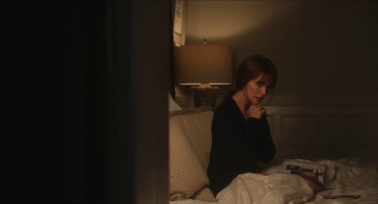 May December (2023) movie still: medium — A dimly lit bedroom features a beige headboard and a warm table lamp. A woman sits on the bed edge while…