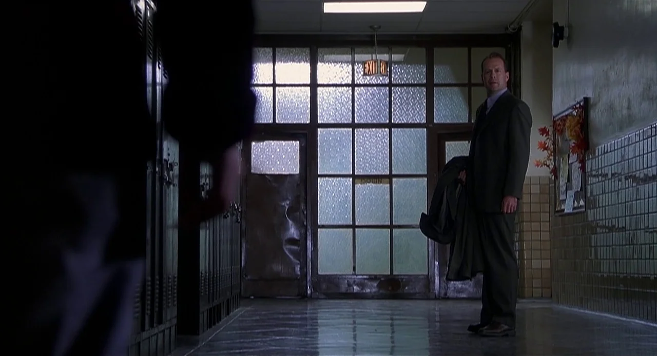 The Sixth Sense (1999) movie still: wide — A school hallway features rows of metal lockers and tiled walls leading to glass doors. A man in a suit…