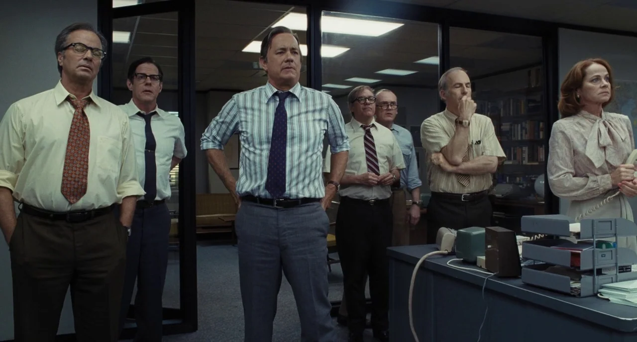 The Post (2017) movie still: medium — A 1970s newspaper office with glass partitions and beige filing cabinets. A group of professionals standing…