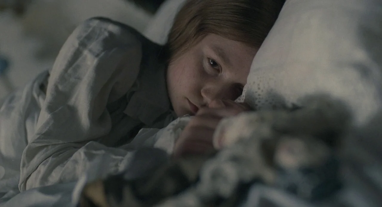 Jane Eyre (2011) movie still: closeup — A child rests in a dim bed with white sheets and pillows. She lies on her side holding a small doll against…